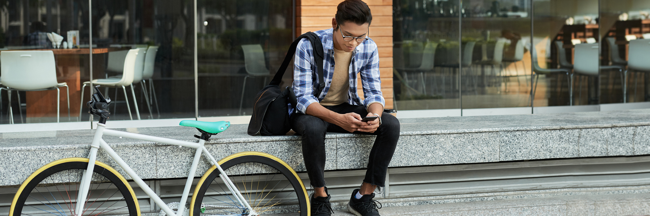 Portrait of cyclist sitting in front of modern coffee house and browsing Internet on smartphone.