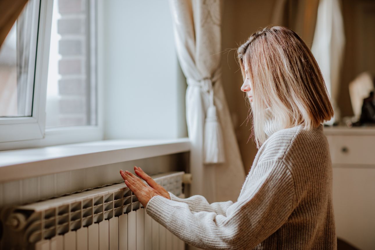 Consumer nz website promo image  young blond woman in long winter beige sweater is warming herself near a heater width