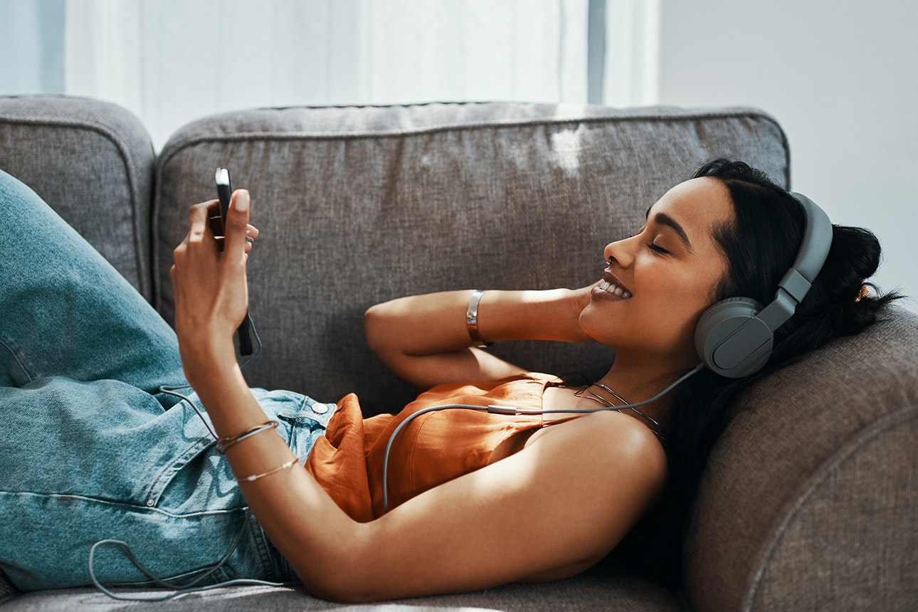 Shot of a young woman using a smartphone and headphones on the sofa at home